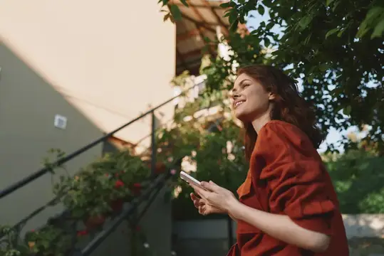 Непринужденный портрет на открытом воздухе улыбающейся женщины в красной одежде, пользующейся телефоном, расслабленная поза, естественный свет, садовая обстановка, лайфстайл-момент