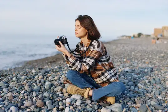 Молодая женщина с фотокамерой сидит на каменистом берегу у моря