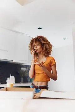 Кудрявая девушка ест пиццу и говорит по телефону на светлой кухне