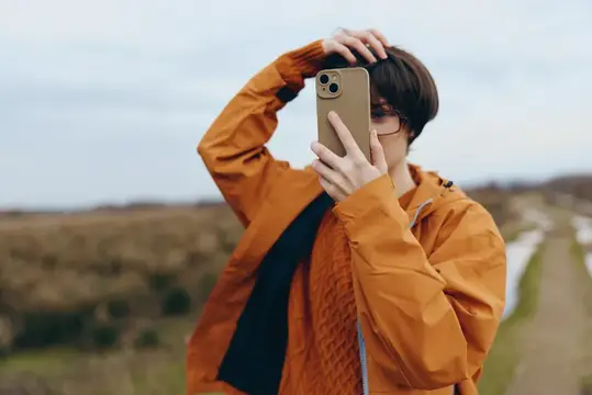 Молодой человек в оранжевой куртке фотографирует на смартфон на фоне поля