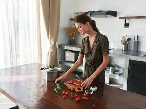 Женщина с улыбкой на кухне готовит обед из мяса, овощей и специй на деревянном столе на фоне стильной кухни