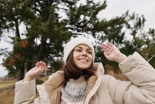 Улыбающаяся девушка в белой шапке и пуховике среди сосен