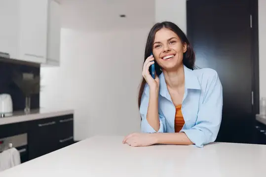 Улыбающаяся темноволосая женщина говорит по телефону на светлой кухне