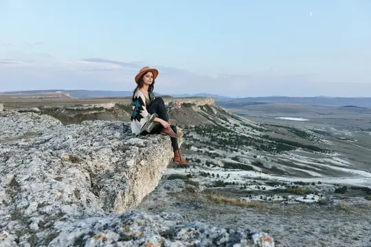 Девушка в шляпе сидит на скале над долиной