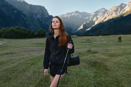 Девушка с рюкзаком стоит на лугу на фоне гор