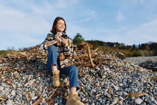 Улыбающаяся девушка с фотоаппаратом сидит на каменистом берегу
