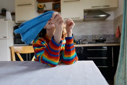 Рыжеволосый мальчик в полосатой кофте играет с синей тканью на кухне