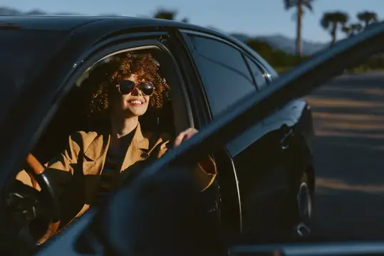 Улыбающаяся кудрявая девушка в солнцезащитных очках за рулём автомобиля