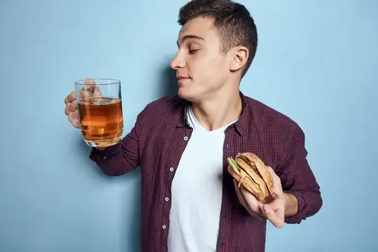 Весёлый пьяный мужчина с кружкой пива и гамбургером в руке, диетическая еда, образ жизни, синий фон