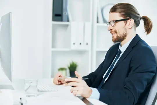 Бородатый мужчина с хвостом и в очках улыбается за компьютером в офисе