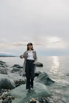 Молодая женщина в плаще и кепке стоит на камнях у моря