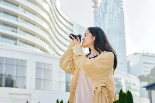 Азиатская девушка фотографирует на плёночную камеру на фоне городских зданий