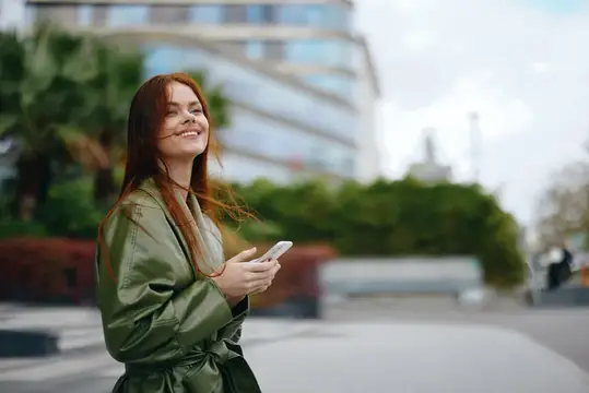 Рыжеволосая девушка со смартфоном улыбается на городской улице