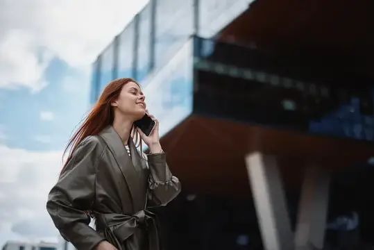 Рыжеволосая девушка в плаще улыбается, слушая телефон у современного здания