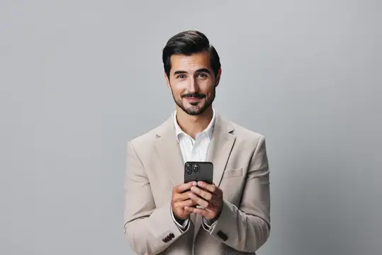 Улыбающийся мужчина в бежевом пиджаке держит смартфон на сером фоне