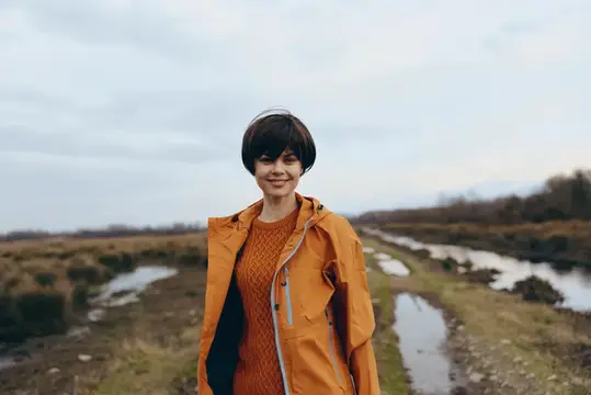 Улыбающаяся темноволосая женщина в оранжевой куртке на фоне осенней степи