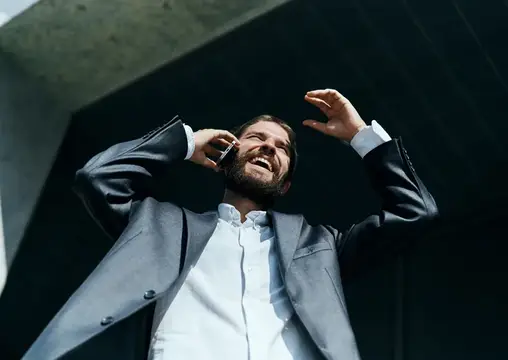 Улыбающийся бородатый мужчина в костюме говорит по телефону на фоне тёмного здания