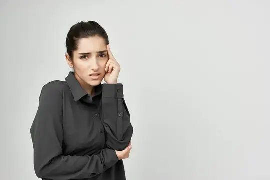 Уставшая темноволосая женщина в серой рубашке держится за голову