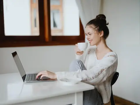 Улыбающаяся девушка с чашкой кофе работает за ноутбуком у окна