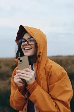 Отдых на природе: женщина в оранжевой куртке с капюшоном держит смартфон, улыбается с радостным выражением лица, свежий воздух природы, мягкий свет облаков, спокойные загородные поля, расслабленная…