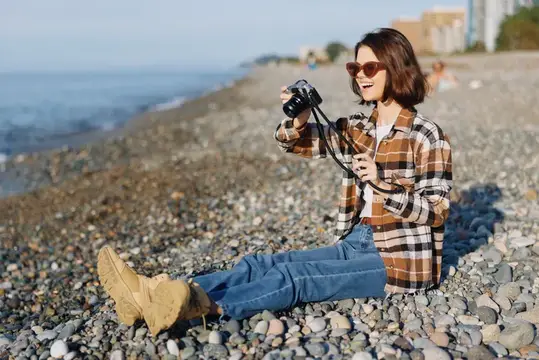 Девушка в клетчатой рубашке сидит на галечном пляже с фотоаппаратом