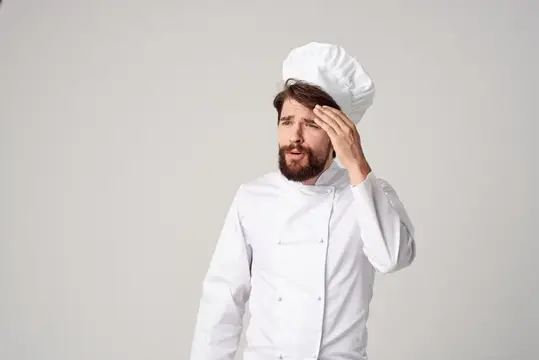 Уставший бородатый повар в белом колпаке вытирает лоб