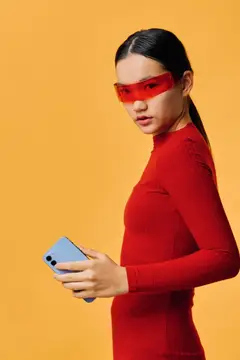Модная женщина в красном платье и красных очках держит смартфон, позируя в студии