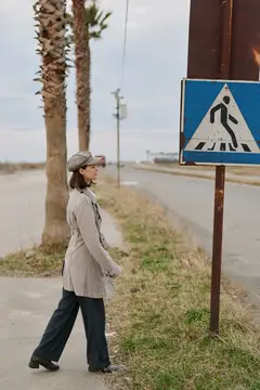 Женщина в кепке и плаще идёт мимо знака пешеходного перехода