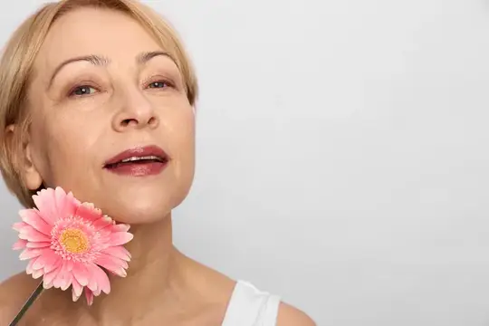 Светловолосая женщина с розовой герберой у лица на сером фоне