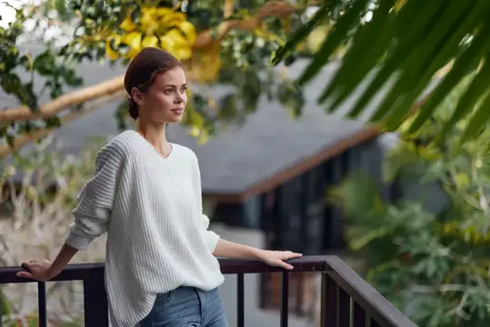 Молодая женщина в белом свитере стоит на балконе в тропиках