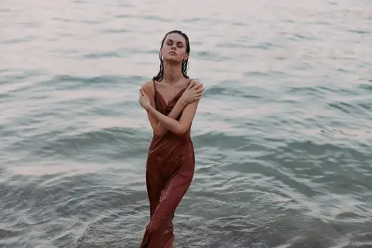 Молодая женщина в мокром платье стоит в воде и обнимает себя