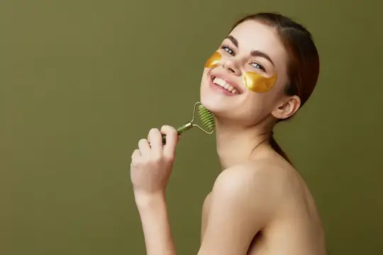 Улыбающаяся девушка с патчами под глазами делает массаж лица роликом