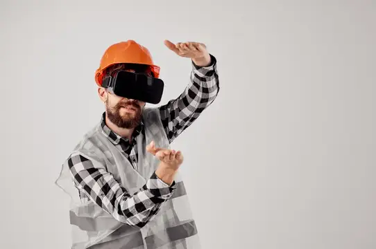 Бородатый мужчина в каске и VR-очках жестикулирует на сером фоне