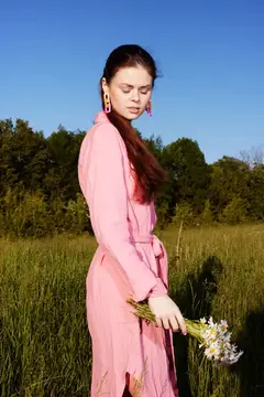 Молодая женщина с длинными волосами в розовом платье стоит на пышном зеленом лугу, держа цветы