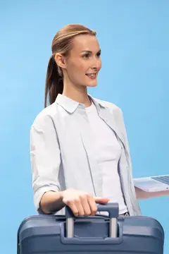 Улыбающаяся женщина в белой рубашке держит ручку чемодана и ноутбук, готовая к путешествию
