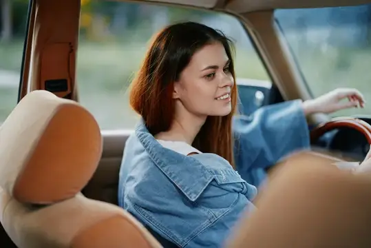 Молодая женщина в модной джинсовой куртке сияюще улыбается за рулем, воплощая чувство свободы и приключений