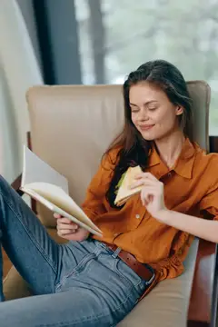 Молодая женщина с сэндвичем читает книгу в кресле