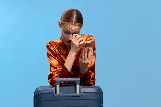Расстроенная женщина в оранжевой блузке вытирает слезы, держа смартфон и опираясь на синий чемодан