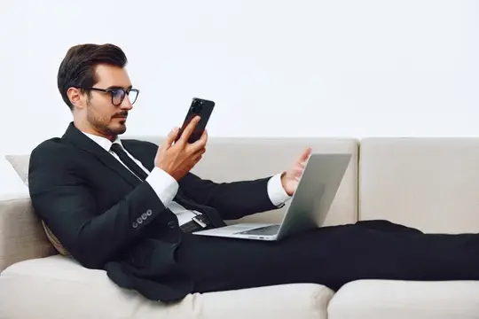 Мужчина-бизнесмен в современном офисе улыбается, используя смартфон и ноутбук