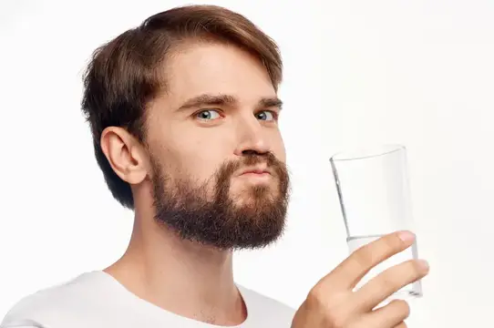 Молодой бородатый мужчина смотрит на стакан воды на белом фоне