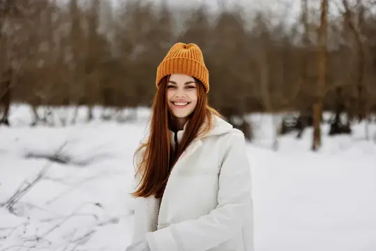 Улыбающаяся рыжеволосая девушка в оранжевой шапке зимой в лесу