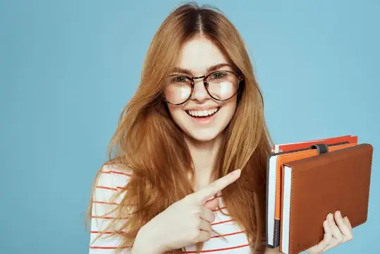 Рыжеволосая девушка в очках улыбается и указывает на стопку книг