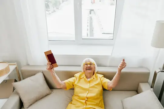 Седовласая женщина в жёлтой рубашке радостно читает книгу на диване