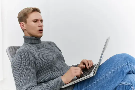 Повседневно одетый молодой человек со светлыми волосами работает за серебристым ноутбуком, на нем серая водолазка, он удобно сидит на однотонном белом фоне, демонстрируя использование технологий и…
