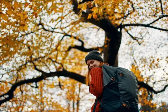 Улыбающаяся девушка в шапке с рюкзаком осенью под деревом