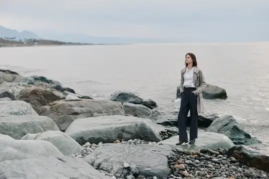 Молодая женщина в плаще стоит на камнях у моря в пасмурный день