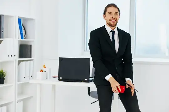 Улыбающийся мужчина в костюме опирается на стол в светлом офисе