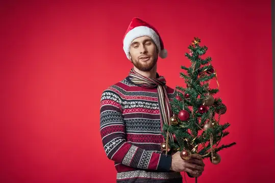 Молодой мужчина в новогоднем свитере и шапке Санты с ёлочкой на красном фоне