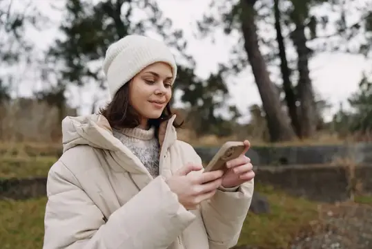 Молодая женщина в толстой куртке и вязаной шапке на улице в парке, листает телефон