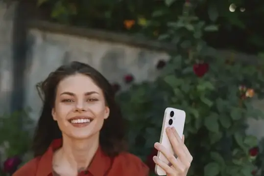Улыбающаяся темноволосая женщина с телефоном на фоне розовых кустов
